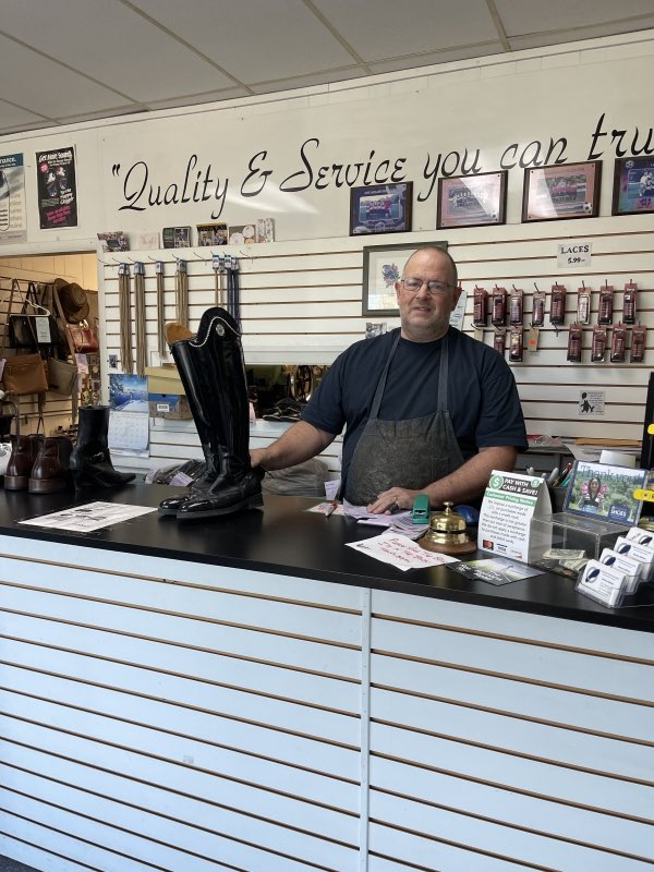 Owner Bryan standing at the shop counter holding a tall repaired black riding boot